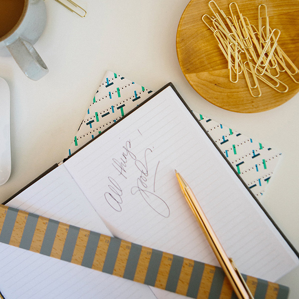 Downward photo of a notebook, gold pen, coffee cup and paperclips on a white desk.